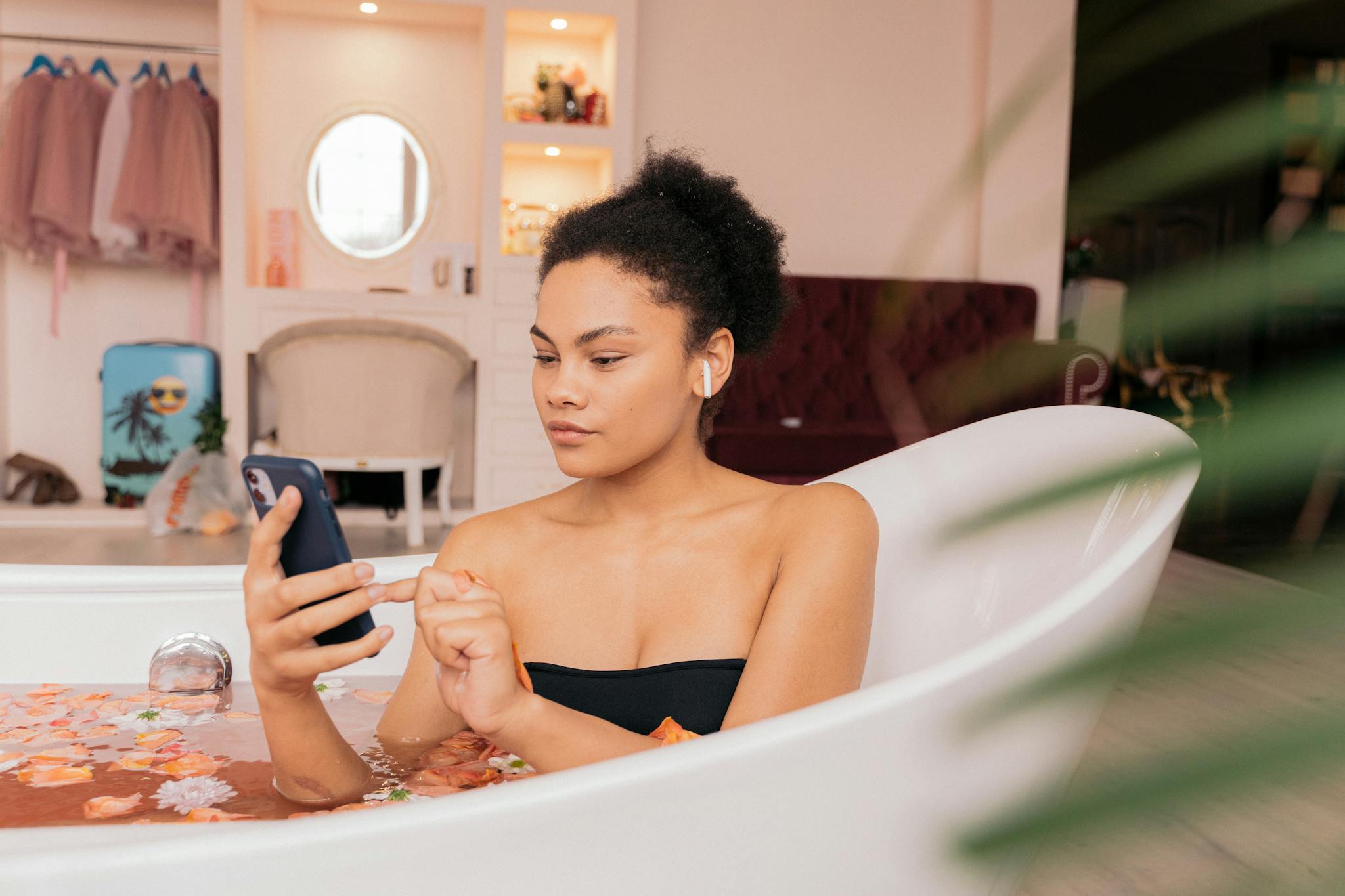 A woman enjoying a relaxing bath with flowers, using a smartphone, indoors.
