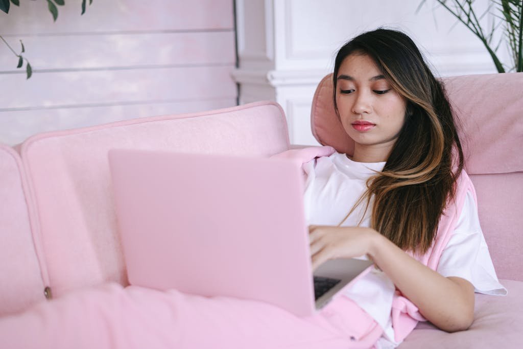 Asian woman in white t-shirt lounging on a pink couch using laptop indoors.