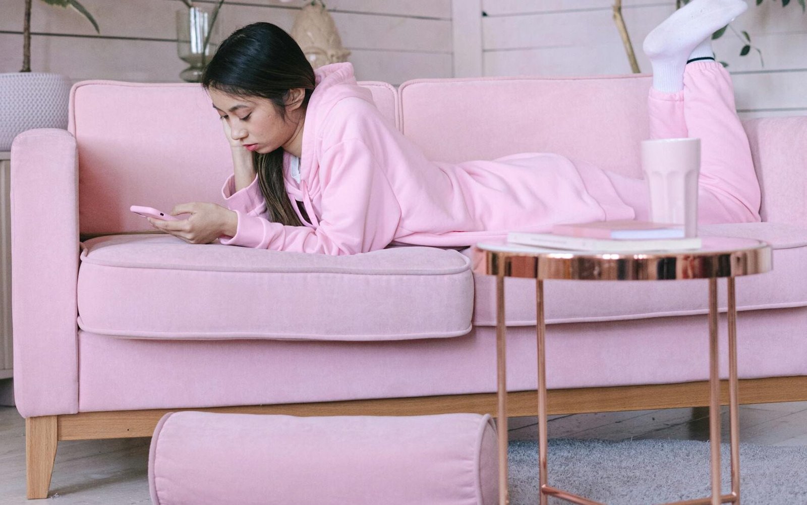 Young woman relaxing on a pink sofa using her smartphone, surrounded by plants and decor.